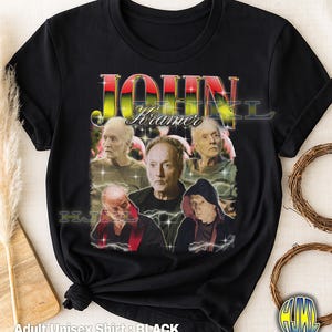Könnte beinhalten: Schwarzes Unisex-T-Shirt für Erwachsene mit einer Grafik von John Kramer aus der Saw-Filmreihe. Das Design zeigt mehrere Bilder der Figur mit dem Namen "John Kramer" in einer farbenfrohen, stilisierten Schriftart. Das T-Shirt ist unten geknotet.