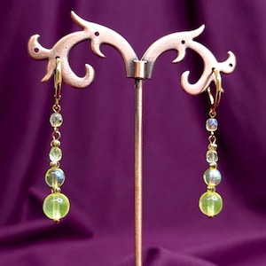 Peut inclure: Une paire de boucles d'oreilles pendantes avec des crochets dormeuses dorés et une disposition verticale de perles. Chaque boucle comprend de petites perles facettées transparentes, des perles sphériques irisées et des perles vert citron.