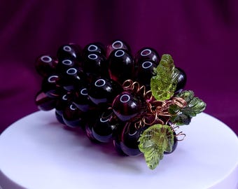 Puede incluir: Un racimo decorativo de uvas de vidrio de color púrpura oscuro con cuentas esféricas translúcidas de color violeta intenso. Las uvas están acentuadas con hojas de vidrio texturizado de color verde brillante y tallos de alambre de cobre.