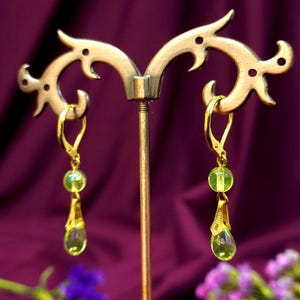 Pode incluir: Um par de brincos dourados com fecho de alavanca, com contas verdes e pingentes em forma de gota. Os brincos são exibidos em um suporte dourado decorativo, contra um fundo roxo. Design delicado e elegante.