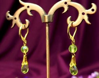May include: A pair of gold-colored leverback earrings with green beads and teardrop-shaped pendants. The earrings are displayed on a decorative gold-colored stand against a purple background. The earrings have a delicate and elegant design.