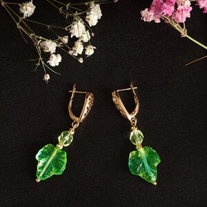 Pode incluir: Um par de brincos pendurados dourados com um design de folha. Cada brinco apresenta um pingente em forma de folha verde e duas contas redondas transparentes. Os brincos estão em um fundo escuro com flores brancas e rosas.