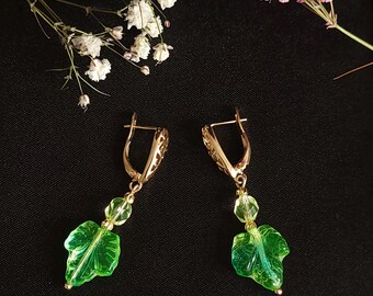 May include: A pair of gold-colored dangle earrings with a leaf design. Each earring features a green leaf-shaped charm and two clear, round beads. The earrings are set against a dark background with white and pink flowers.