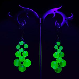 Pode incluir: Um par de brincos pendentes com cachos de contas redondas verde-neon que brilham sob luz ultravioleta. Cada brinco está suspenso por um gancho de metal prateado e consiste em várias contas esféricas numa formação em cascata.