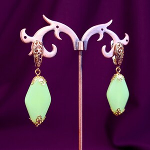 Pode incluir: Um par de brincos dourados com pedras verde-claras em formato de diamante. Cada brinco possui uma tampa de metal dourado com filigrana nas extremidades, suspenso por um fecho de alavanca a condizer e ornamentado.