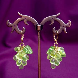 Pode incluir: Um par de brincos pendentes com cachos de contas de vidro verde-claro translúcido em forma de uva, acentuados com uma folha de vidro verde em cada um. Os brincos estão suspensos em ganchos de alavanca de metal dourado.