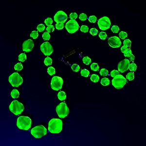 May include: A vibrant, neon green necklace featuring faceted beads. The necklace is made up of multiple strands of varying bead sizes, with a clasp at the back. The beads glow brightly against a dark backdrop, creating a striking visual effect. A statement necklace.