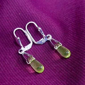 Pode incluir: Um par de brincos prateados com fecho de alavanca, apresentando pequenos pingentes de vidro translúcido em forma de lágrima na cor verde-clara. As peças metálicas são polidas e prendem as gotas de vidro lisas e arredondadas.