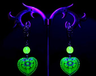 May include: A pair of neon green heart-shaped earrings with a floral design, suspended from a decorative stand. Each earring features a round bead and intricate detailing, glowing brightly against a black background. The earrings are designed to stand out.