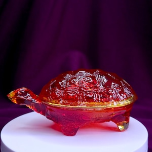 May include: A translucent red glass turtle figurine with a detailed shell pattern. The object features a molded head, four short legs, and a rounded back with geometric textures. The glass displays varying shades of deep red and orange light.