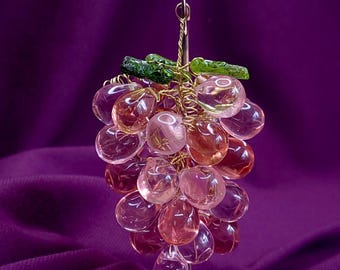 May include: A decorative cluster of translucent pink glass beads shaped like grapes, featuring two textured green glass leaves at the top. The beads are connected by thin, twisted gold-toned wire, forming a bunch with a small metal loop for hanging.