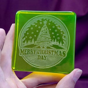 以下が含まれることがあります： 雪に囲まれたクリスマスツリーの円形デザインがエッチングされた、半透明のライムグリーンの正方形のガラス製コースター。円の下部には、太字のセリフ体で「MERRY CHRISTMAS DAY」という文字が刻印されています。