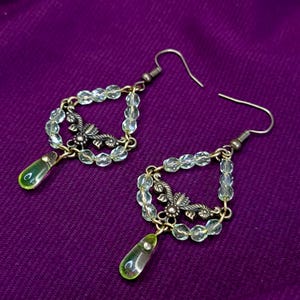 Può includere: Un paio di orecchini pendenti con una montatura a goccia composta da piccole perline sfaccettate trasparenti. Ogni orecchino include un elemento centrale floreale in metallo color bronzo antico e una perla di vetro verde chiaro.