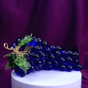 May include: A decorative cluster of deep blue glass grapes featuring translucent green glass leaves and a gold-toned wire stem. The individual glass grapes are smooth and rounded, arranged in a dense bunch with reflective highlights on their surfaces.