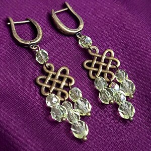 Pode incluir: Um par de brincos pendentes com ganchos de alavanca em metal tom bronze antigo e um design central de nó celta. Cada brinco é adornado com múltiplas contas de cristal facetado em verde lima pálido suspensas do elemento de nó.