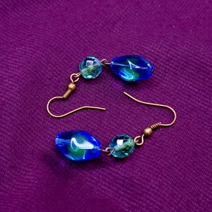 Pode incluir: Um par de brincos pendentes com ganchos de metal em tom de bronze. Cada brinco consiste numa pequena conta facetada de cor azul-petróleo translúcida combinada com uma conta de vidro azul maior, lisa e irregular, com subtis reflexos verdes e brancos.