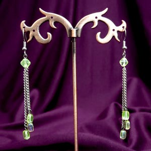 Pode incluir: Um par de brincos pendentes com correntes de metal prateado e contas verde-claras translúcidas. Cada brinco tem uma conta facetada no topo, seguida por duas correntes prateadas paralelas que terminam em três pequenas contas verdes retangulares.