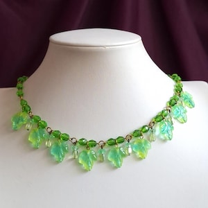 May include: A green beaded necklace with leaf-shaped pendants. The necklace features small green beads and larger, translucent green and yellow leaf charms. The necklace is displayed on a white mannequin.