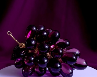 May include: A decorative cluster of translucent, deep purple glass grapes arranged in a bunch. The individual grapes are oval-shaped and connected by thin, twisted gold-toned wire, with a small wire loop extending from the top of the cluster.