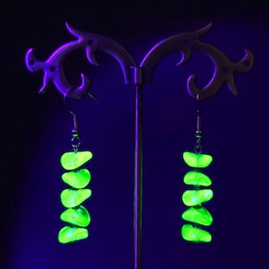 Pode incluir: Um par de brincos pendentes com cinco pedras irregulares empilhadas de cor verde néon brilhante, suspensas por ganchos de metal prateado. As pedras emitem uma luz intensa contra um fundo escuro, criando um efeito de alto contraste.