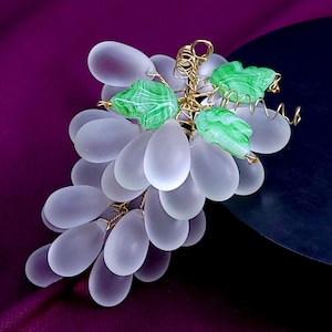 Op de afbeelding: Een decoratieve tros van mat, doorschijnend wit glas in de vorm van druiven. De druiven zijn verbonden door dun, gedraaid goudkleurig draad, dat ook drie kleine, gesneden groene glazen bladeren met witte nerven bevat.