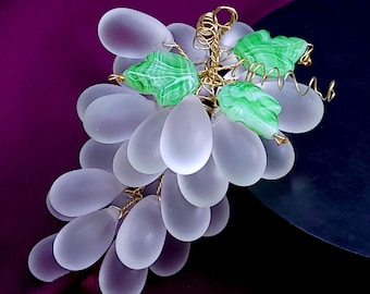 May include: A decorative cluster of frosted, translucent white glass grapes arranged in a bunch. The grapes are connected by thin, twisted gold-toned wire, which also features three small, carved green glass leaves with white veining.
