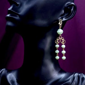 Pode incluir: Um brinco pendente de tom dourado com uma conta verde-pálida no topo, seguido por um conector decorativo de filigrana dourada que se ramifica em duas correntes separadas, cada uma adornada com três contas verde-pálidas adicionais pendentes.