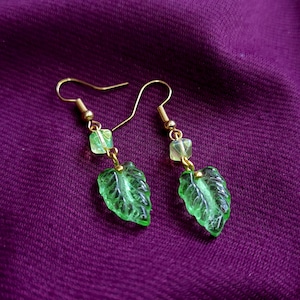 Pode incluir: Um par de brincos pendentes com ganchos de metal dourado, cada um ligado a uma pequena conta de cubo verde-claro translúcido e a um pendente de vidro verde em forma de folha com nervuras detalhadas e acabamento.