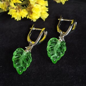 Pode incluir: Um par de brincos de alavanca prateados com pingentes de folhas verdes translúcidas. As folhas têm veias detalhadas e são suspensas dos aros prateados. Flores amarelas no fundo.