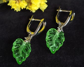 May include: A pair of silver-tone leverback earrings with translucent green leaf pendants. The leaves have detailed veining and are suspended from the silver hoops. Yellow flowers are in the background.