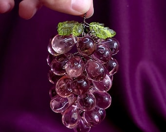 Puede incluir: Un racimo decorativo de uvas de vidrio púrpura translúcido con dos hojas de vidrio verde claro texturizado en la parte superior. Las uvas esféricas individuales están conectadas por un fino alambre de metal oscuro, creando un racimo de frutas realista.
