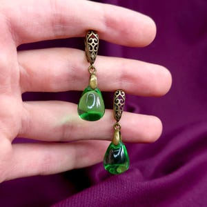 Pode incluir: Um par de brincos pendentes com contas de vidro translúcido em forma de lágrima verde, suspensas por ganchos de metal bronze com filigrana ornamentada de estilo antigo. Os brincos têm um acabamento liso e polido.