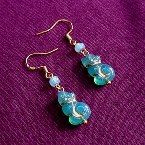 Pode incluir: Um par de brincos de gancho em tom dourado com uma pequena conta azul-clara e um pingente translúcido em forma de gato na cor verde-azulado com detalhes dourados. Os pingentes de gato têm uma cauda curva e um acabamento liso e polido.