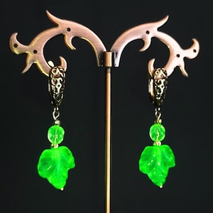 Pode incluir: Brincos pendurados com um design de folha. Os brincos apresentam uma parte superior de filigrana dourada, uma pequena conta verde e um pingente em forma de folha verde vibrante. São exibidos em um suporte decorativo de cor cobre sobre um fundo escuro.