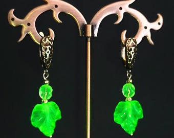 May include: A pair of dangle earrings with a leaf design. The earrings feature a gold-colored filigree top, a small green bead, and a vibrant green leaf-shaped charm. The earrings are displayed on a decorative copper-colored stand against a dark background.