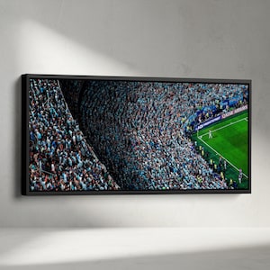 May include: A panoramic framed wall art print depicting a high-angle view of a crowded soccer stadium. The stands are filled with spectators wearing light blue, contrasting with the bright green grass of the pitch and visible stadium signage.