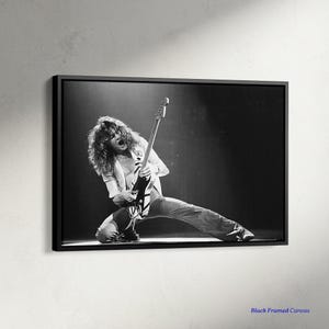 May include: Black and white framed canvas print of a musician playing a guitar on stage. The musician is kneeling, wearing a white shirt and light-colored pants. The guitar has a black body with white stripes. The image is set against a dark background.