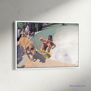 May include: A framed canvas print featuring a vintage skateboarding scene. A skater in a black hat rides a yellow skateboard in a pool, with a dog nearby. The print is in a white frame, capturing a moment of action and leisure.