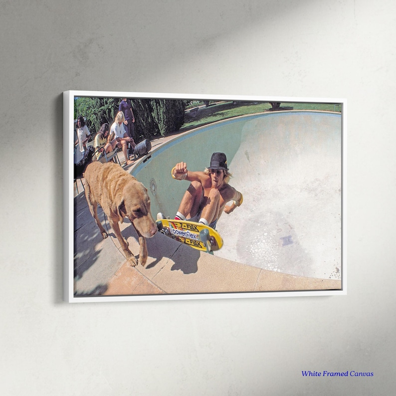 May include: A framed canvas print featuring a vintage skateboarding scene. A skater in a black hat rides a yellow skateboard in a pool, with a dog nearby. The print is in a white frame, capturing a moment of action and leisure.