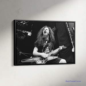 May include: Black and white framed canvas print of a musician playing a guitar on stage. The musician is wearing a black t-shirt and shorts, with long hair and a necklace. The guitar is a V-shaped model. The canvas is framed in black.
