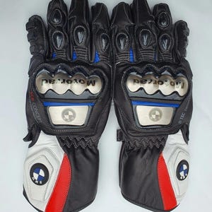 K&ouml;nnte beinhalten: Paar schwarze und wei&szlig;e Motorradhandschuhe mit roten und blauen Akzenten. Die Handschuhe zeigen das BMW-Logo und das Wort "Motorrad". Sie verf&uuml;gen &uuml;ber Schutzpolster und N&auml;hte.