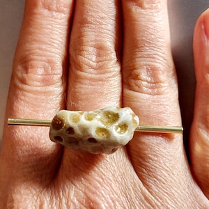 May include: A unique ring featuring a porous, beige-colored stone with brown spots, mounted on a thin, gold-colored band. The stone has a textured surface and is the focal point of the jewelry piece.