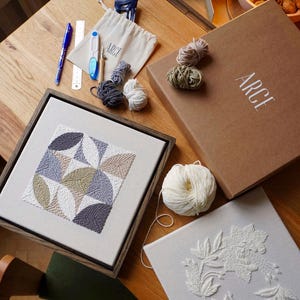 Könnte beinhalten: Auf einem Holztisch liegen Stickutensilien und fertige Stickereien. Dazu gehören ein gerahmtes Stickbild mit geometrischem Muster, eine Box mit dem Wort "ARCE", Garn und ein zweites Stickbild mit floralem Design.
