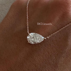 May include: A delicate gold necklace featuring a pear-shaped diamond pendant. The diamond is clear and sparkling, suspended from a fine chain. The necklace is displayed on a hand, showcasing the jewelry's elegance and design.