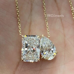 May include: A gold necklace featuring two diamond pendants: a rectangular cut and a pear-shaped cut. The diamonds sparkle with multiple facets. The necklace is held in a hand, with the brand name "RKD Jewels" visible in the background.
