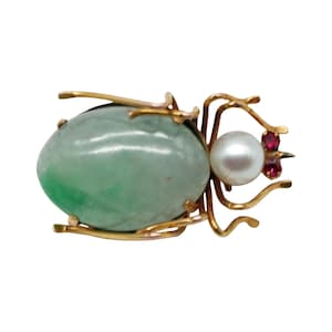 May include: A gold-toned insect brooch featuring a large, oval, pale green jade body. The brooch has delicate gold legs, a white pearl, and two small red gemstones. The design is intricate and detailed.