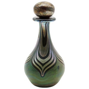 May include: A decorative glass perfume bottle with a silver-toned stopper. The bottle has a teardrop shape with a green and iridescent finish, featuring a black and silver striped pattern. The stopper is a rounded shape with a metallic finish.