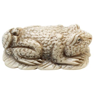 May include: A detailed ivory-colored frog figurine with a textured surface and dark spots. A small turtle rests on the frog's back. The frog is positioned on a leaf-like base, showcasing intricate carving details. The figurine is a decorative item.