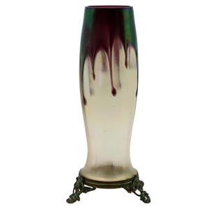 May include: A tall, iridescent glass vase with a flared base and a dark, dripped design. The vase has a bronze-colored metal base with ornate, curved legs. The top of the vase is a deep burgundy color that fades into a lighter, pearlescent hue.