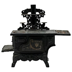 May include: A black antique cast iron stove with ornate detailing. The stove features the word "RESCENT" and a star design on the front. It has a decorative back piece and a side extension. The stove is a vintage kitchen appliance.